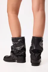 Bota de caña media estilo slouchy en color negro, confeccionada en material sintético con acabado liso. Presenta un diseño holgado en la caña, puntera redondeada, suela plana y una tira decorativa con hebilla metálica ajustable en la zona del tobillo.