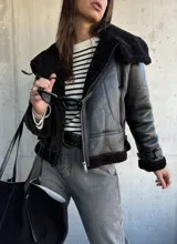 Campera tipo gamulán de cuero sintético negro con interior y detalles de cuello y puños en piel sintética negra. Presenta cierre frontal, correas ajustables en los puños y diseño de corte clásico.
