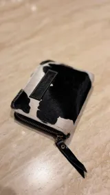 Billetera de cuero de pelo con estampado animal print tipo vaca en blanco y negro. Cuenta con cierre perimetral y tirador de cuero con detalle metálico.