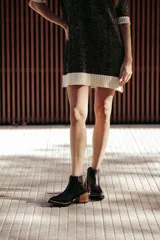 Botín de cuero negro, caña corta, con punta cuadrada y cierre lateral. Presenta un tacón de madera de altura media y detalles en charol con textura de cocodrilo en el talón.