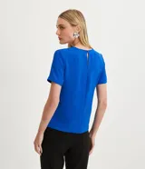 Blusa blanca de poliéster con cuello redondo, mangas cortas, cierre trasero en forma de gota, pequeñas aberturas laterales y acabados en pespuntes en las mangas y el cuello.