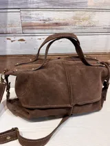Bolso de mano tipo bandolera confeccionado en cuero gamuzado color marrón. Presenta doble asa superior, correa larga ajustable y extraíble, cierre superior con cremallera y detalles de remaches metálicos en color bronce.