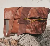 Bolso tipo bandolera de cuero color marrón, con diseño de solapa plegable y detalle de tira central en cuero más oscuro con logo grabado. Presenta una correa larga ajustable y una silueta amplia y versátil.