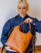 Bolso tipo matera de cuero con textura de reptil, color tostado, con manijas trenzadas y detalles de costura artesanal en los bordes.