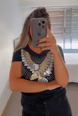 Remera blanca de algodón con cuello redondo y manga corta. Presenta un bordado de un águila con lentejuelas doradas, plateadas y negras en el frente.