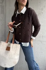 Cardigan de punto color chocolate, con diseño de trenzas y pompones decorativos. Presenta cuello redondo, cierre frontal con botones metálicos dorados y mangas largas.