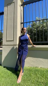 Vestido largo de fiesta color azul, con cuello redondo, sin mangas y diseño ajustado. Presenta un detalle de nudo en la cintura con una abertura frontal y una falda superpuesta de tul transparente.