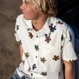 Camisa de manga corta color blanco con estampado de flores pequeñas en tonos oscuros y tierra. Presenta cuello clásico, cierre frontal con botones y un bolsillo en el pecho.
