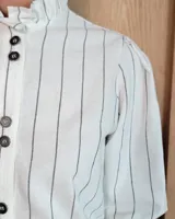 Camisa de algodón a rayas verticales blancas y azules, con cuello alto tipo gorguera con volados, cierre frontal de botones y mangas largas con puños con volados.