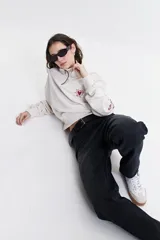 Buzo de felpa color crudo con cuello alto, cierre metálico y bordado de un corazón con rayos en el pecho.