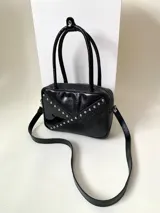 Cartera rígida negra con detalles en rojo y tachas plateadas. Tiene herrajes y cierre metálico plateado marca YKK, asa fina para llevar al hombro o en la mano y correa larga extra regulable. El interior es forrado, tiene un bolsillo con cierre enmarcado en cuero, y un segundo bolsillo plaqué.