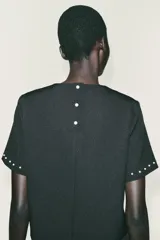 Top negro de manga corta con cuello redondo, adornado con perlas blancas aplicadas en el escote y en los puños de las mangas. Cierre posterior con botones de perla.