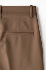 Pantalón tobillero color marrón, de tejido elástico, con cintura estándar y elástico oculto. Presenta cierre con corchete oculto, bolsillos al bies y bolsillos traseros decorativos. Las piernas son ajustadas.