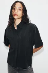 Camisa de manga corta color marrón oscuro, con cuello solapa, cierre frontal oculto con botones y dos bolsillos de parche en el pecho.
