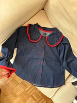 Camisa de jean azul oscuro con cuello estilo marinero y puños con volados, ambos con ribetes en color rojo. Presenta cierre frontal con botones rojos.