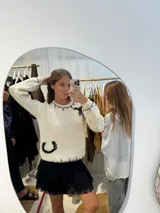 Buzo tejido a mano en color crudo, con diseño de herraduras bordadas en contraste negro y detalles de pespuntes en cuello, hombros y puños. Presenta un corte clásico de cuello redondo y mangas largas.