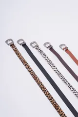 Cinturón de estilo western con hebilla metálica plateada ornamentada. Disponible en varios colores y estampados: cuero marrón claro, cuero marrón oscuro, estampado dálmata (blanco y negro) y cuero negro liso. También disponible en estampado leopardo.