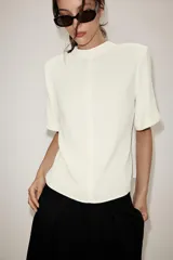 Camisa de cuello subido y manga corta con hombreras. Presenta un diseño minimalista con cierre de cremallera metálica en la espalda.
