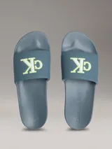 Ojotas tipo slide de Calvin Klein Jeans en color azul, con logo de la marca en relieve en color verde neón sobre la banda superior y logo grabado en el lateral de la suela.