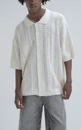 Camisa de manga corta tejida en punto abierto tipo crochet, con cuello polo y cierre frontal de botones. Presenta un diseño oversize y textura calada.