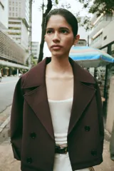 Trench corto de color marrón oscuro, con cuello de solapa, cierre frontal cruzado con botones y mangas tres cuartos con detalle de vuelta, trabilla y botón. Cuenta con bolsillos delanteros de vivo.