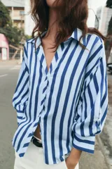 Camisa de rayas verticales azules y celestes, con cuello solapa, escote en V, cierre frontal con botones y mangas largas abullonadas con puños ajustados.