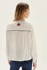 Camisa de algodón color crudo con cuello mao y cierre frontal de botones. Presenta bordados florales en tonos rojo y negro distribuidos en el cuerpo, cuello y puños.