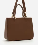 Cartera estilo satchel de tamaño mediano, confeccionada en poliuretano color marrón con textura tipo cuero. Cuenta con asas de mano reforzadas, correa larga desmontable, cierre superior de cremallera y un colgante decorativo de pompón de pelo sintético.