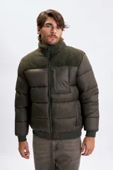 Campera puffer color verde oliva oscuro con diseño capitoneado. Presenta un panel superior en tejido tipo corderito en el pecho y cuello alto. Cuenta con cierre frontal completo, puños y cintura elástica acanalada, y un bolsillo vertical con cierre en el pecho.