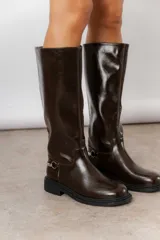 Bota de caña alta con acabado brillante, punta redondeada y suela plana. Presenta un detalle metálico decorativo en el lateral del tobillo.