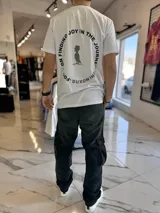 Remera blanca de manga corta con estampa en la espalda que incluye la frase 'Focus on finding joy in the journey' formando un círculo alrededor de la figura de un pájaro con sombrero.