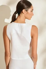 Top de lino blanco sin mangas, con cuello redondo y cierre de botones de madera dispuestos en una cartera asimétrica. Cuenta con delicados detalles bordados en forma de flor alrededor de los ojales.