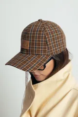 Gorra tipo baseball con estampado de cuadros escoceses en tonos marrones y azules. Presenta un parche rectangular de cuero sintético en el frente con el logo grabado.