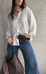 Camisa oversize blanca con botones frontales, cuello camisero y manga larga. Presenta grandes bordados en hilo beige con diseño de hojas o abanicos en la parte inferior frontal.