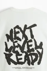 Remera de algodón blanca de corte holgado, con cuello redondo y un pequeño estampado gráfico negro en el pecho con el texto "NEXT LEVEL READY".