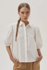 Camisa blanca de mangas abullonadas con puños elásticos, cuello clásico y cierre frontal con botones. Presenta detalles de bordado calado en el pecho.