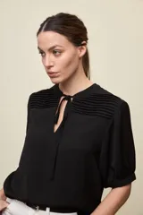 Blusa de manga corta color negro, con calce relajado, escote redondo con abertura y lazo para anudar. Presenta detalles de tablas horizontales en la parte superior que aportan textura al diseño.