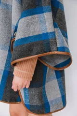 Ruana confeccionada en paño italiano con diseño a cuadros en tonos gris, azul y negro, con terminaciones ribeteadas en cuerina color camel y botón en las mangas.