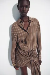 Vestido corto marrón con rayas verticales blancas, cuello camisero, manga larga y detalle de nudo y lazada en la falda. Cuenta con cierre frontal de botones.