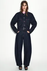 Jeans de tiro alto color azul oscuro, con cinco bolsillos y pernera holgada que se estrecha hacia el tobillo. Presenta pespuntes en color rojo y cierre frontal con cremallera y botón.