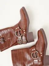 Botas de caña media Mutma modelo Mota, confeccionadas en cuero texturado color marrón. Presentan diseño de punta cuadrada, taco de 5 cm, múltiples tiras con hebillas metálicas ajustables y herrajes circulares en los laterales.