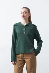 Sweater de tejido de punto color verde oscuro, con cuello polo amplio y cierre frontal parcial con tres botones metalizados. Presenta mangas largas levemente acampanadas y aberturas laterales en el ruedo.