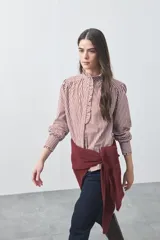 Camisa de manga larga a rayas verticales en tonos blanco y bordó, con cuello alto y detalle de volados frontales. Presenta puños con volados y un corte fluido.