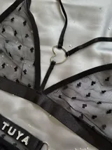 Conjunto de lencería de dos piezas en tul negro con estampado de corazones floca, compuesto por un bralette con tiras ajustables y argolla en forma de corazón, y una tanga con tiras regulables, argollas metálicas y detalle de pedrería con la inscripción 'Soy Tuya'.