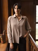 Camisa de color beige con cuello clásico y cierre frontal de botones. Presenta un diseño de bordado lineal en color blanco que recorre las mangas largas.