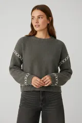 Sweater de tejido de punto grueso color gris oscuro, con cuello redondo a la base y mangas largas. Presenta detalles de bordado decorativo en color blanco con diseño de cruces en las mangas y el ruedo.