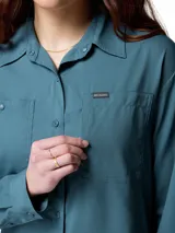 Camisa de senderismo Columbia de manga larga, color azul petróleo, con cuello clásico, cierre frontal con botones y múltiples bolsillos funcionales en el pecho. Presenta tecnología de protección solar y tejido de secado rápido con ventilación trasera.