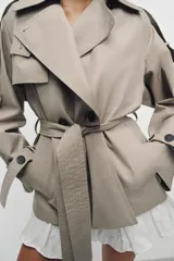 Trench corto de corte oversize confeccionado en mezcla de algodón con acabado repelente al agua. Presenta cuello solapa, cierre frontal con botones, mangas largas con trabillas ajustables y cinturón a tono con lazada en la cintura.