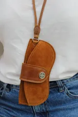 Estuche para lentes con correa larga para colgar al cuello, confeccionado en cuero gamuzado color chocolate. Cuenta con una solapa frontal con cierre de broche metálico a presión.