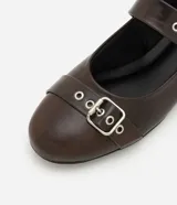 Zapatilla tipo bailarina color marrón oscuro, confeccionada en poliuretano con acabado mate. Presenta punta redondeada, suela plana y dos tiras ajustables con hebillas metálicas plateadas y ojales decorativos sobre el empeine.